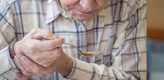 Elderly man with Parkinsons disease holds spoon in both hands.