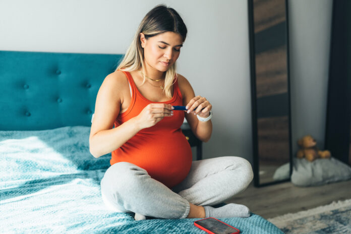 Pregnant young woman sitting on a bed preparing insulin dosage in insulin pen before injecting to represent gestational diabetes and other adverse pregnancy outcomes.