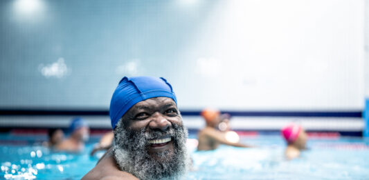 Portrait of an older black man in a swimming pool wearing a swimming hat. Exercise is one of several lifestyle factors that can help increase lifespan.