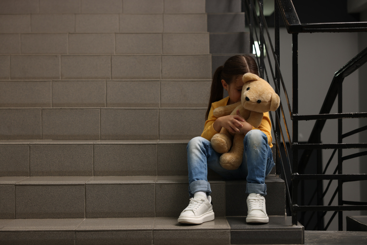 Upset little girl with teddy bear sitting on stairs indoors, space for text